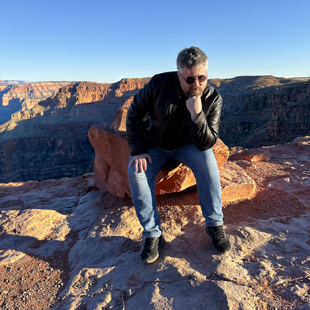 A picture of me wearing sunglasses, sitting on a rock and posing like Rodin's The Thinker in front of the Grand Canyon